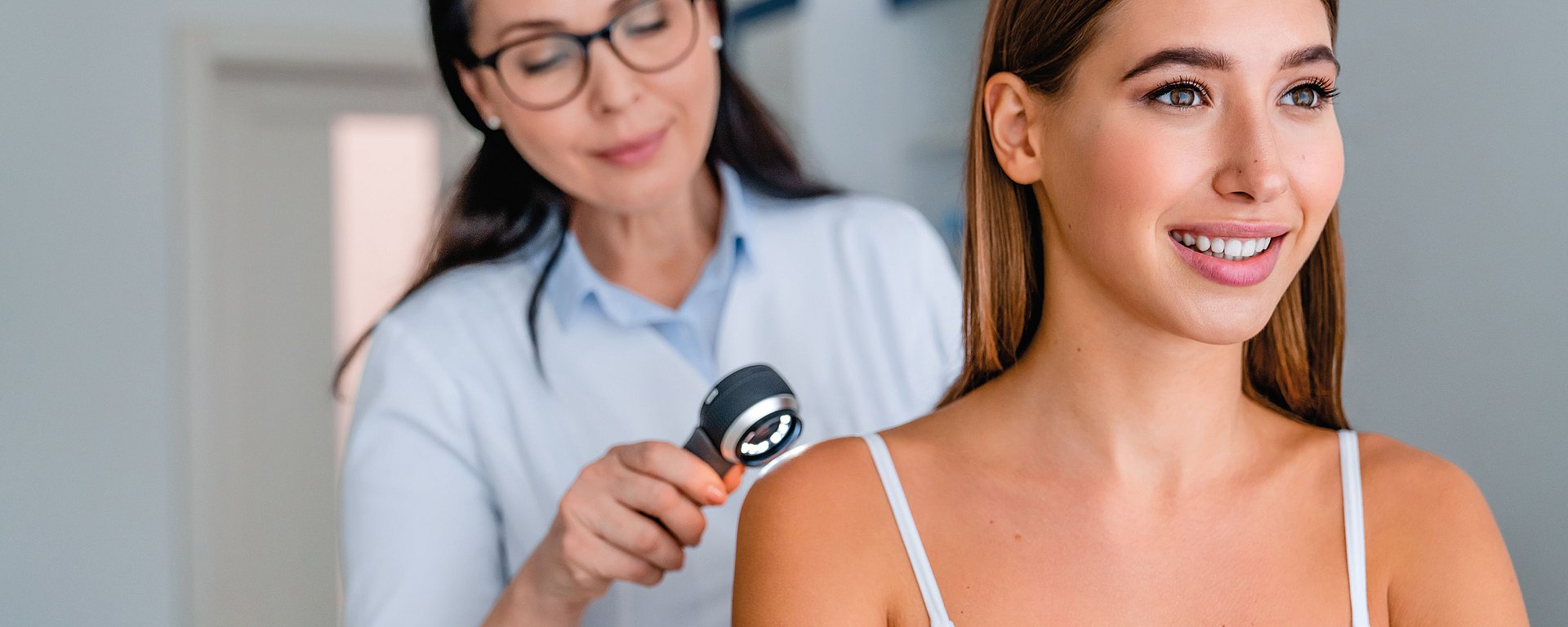 In einer Arztpraxis untersucht ein Arzt die Schulter einer lächelnden Frau mit einem Dermatoskop.
