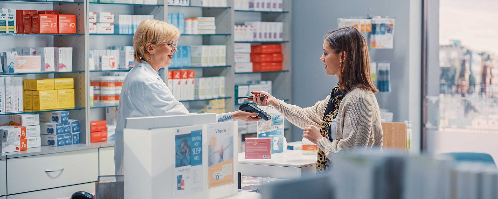 Eine Frau bezahlt einen Apotheker am Apotheken-Schalter, umgeben von Regalen mit Medikamenten und Gesundheitsprodukten.