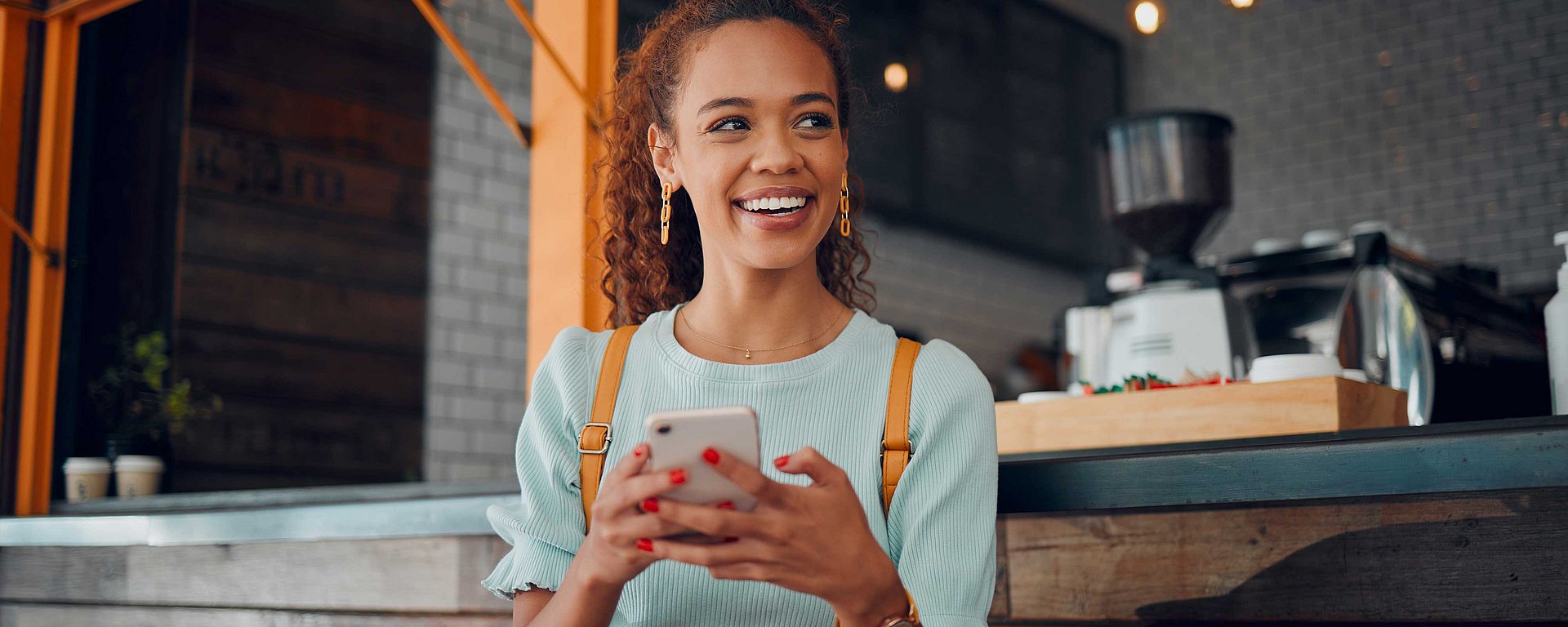 Eine lächelnde Frau, die ein Smartphone in der Hand hält, sitzt in einem modernen Café, im Hintergrund sind Kaffeemaschinen zu sehen.