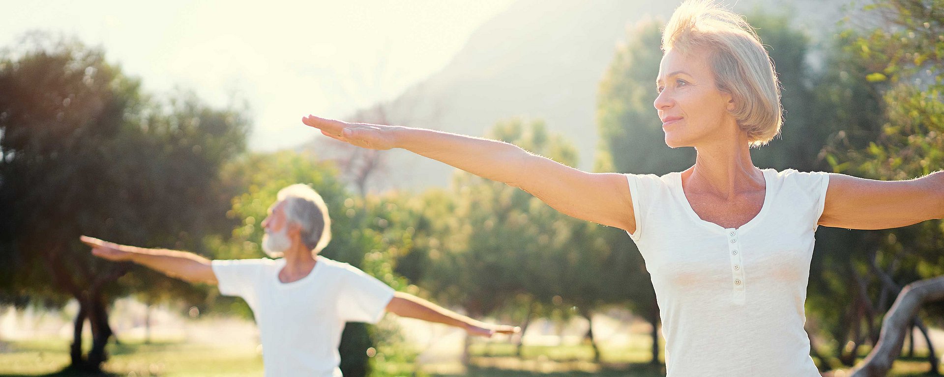 Ältere Menschen, die im Freien Yoga praktizieren, die Arme ausgestreckt, umgeben von Bäumen und Sonnenlicht.