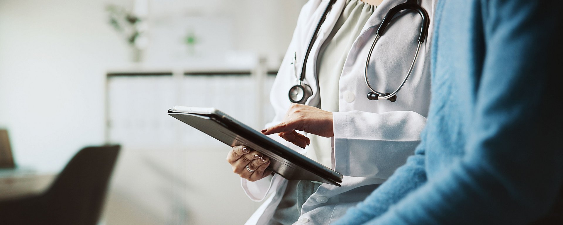 Ein Arzt mit einem Stethoskop nutzt ein Tablet, während er sich in einer Arztpraxis mit einem Patienten unterhält.