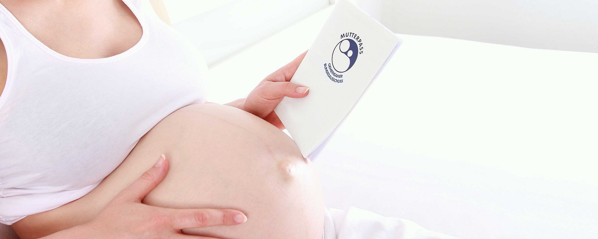 Eine schwangere Person in weißer Kleidung, die ein Schwangerschaftsbuch in der Hand hält, während sie auf einem Bett ruht.