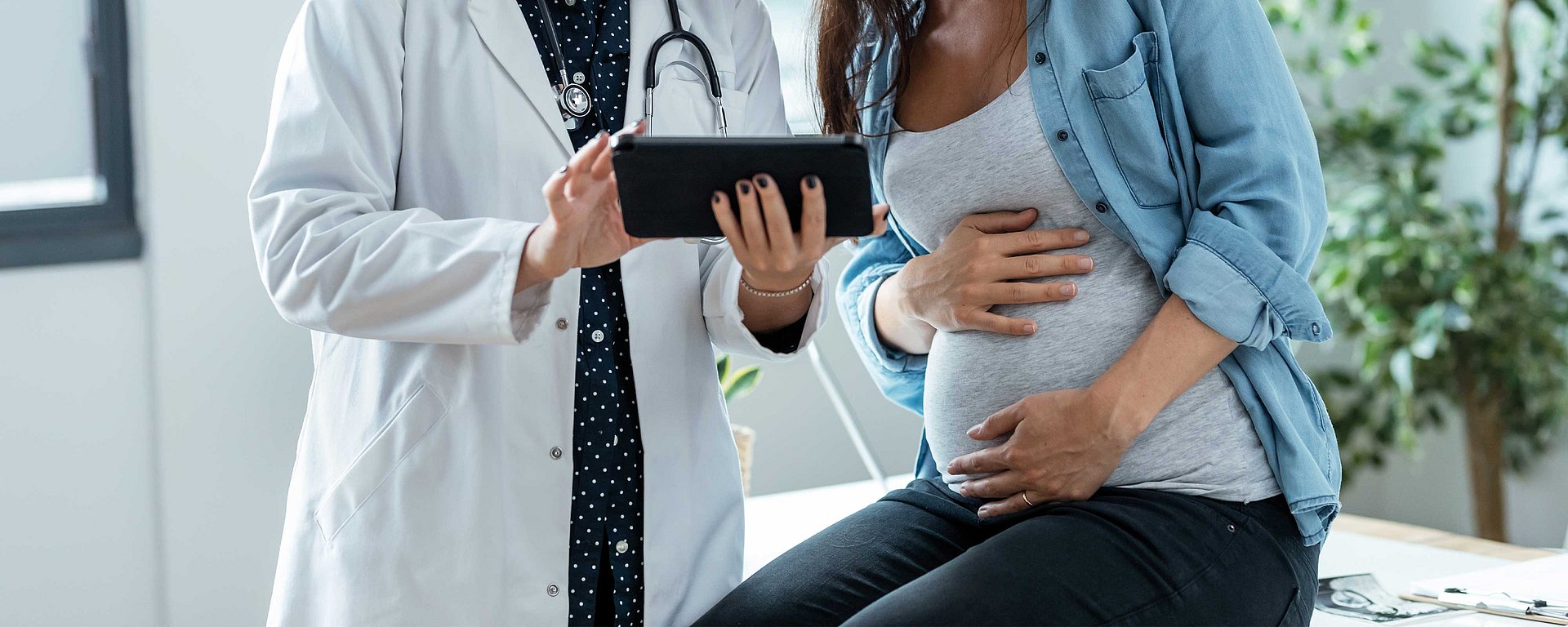 Ein Arzt zeigt einer schwangeren Frau, die während eines Arzttermins ihren Bauch hält, ein Tablet.