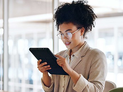Eine Frau mit Brille nutzt ein Tablet, während sie in einem hellen, modernen Büro sitzt.