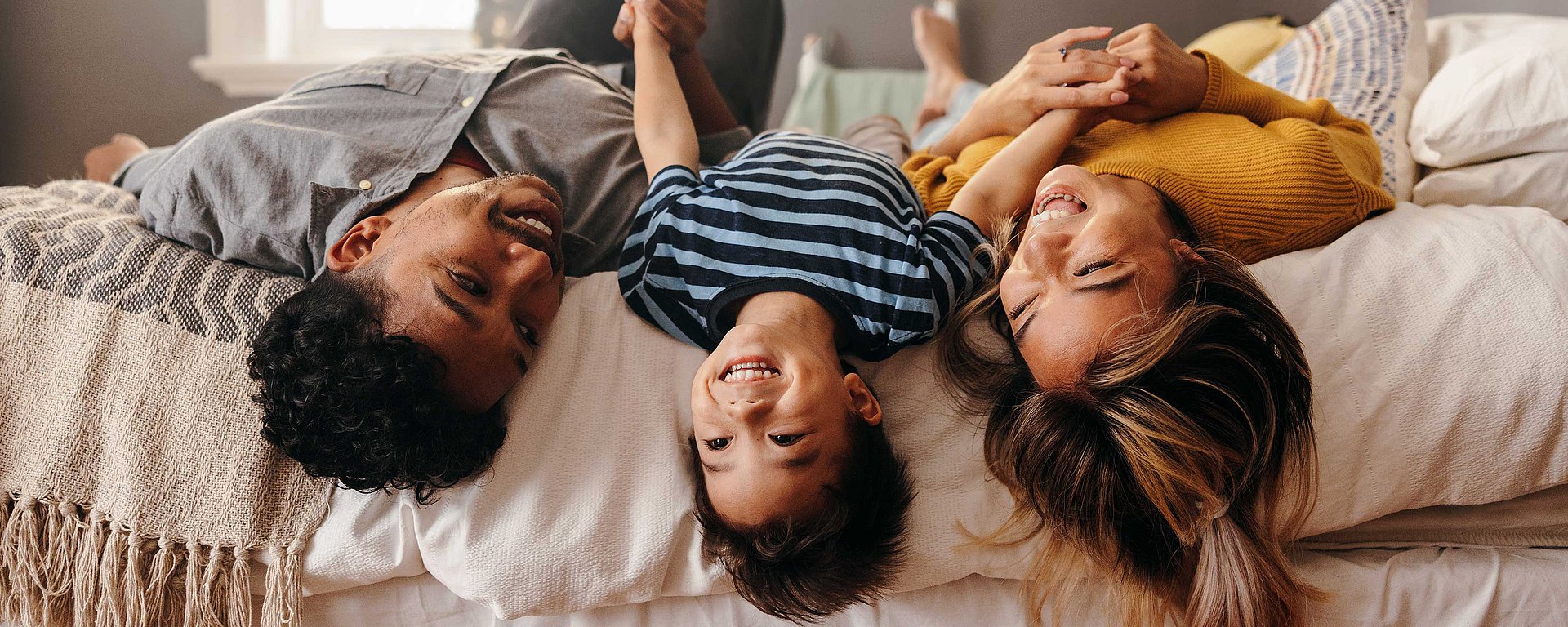 Eine glückliche dreiköpfige Familie liegt auf einem Bett, lächelt und hält sich an den Händen, wobei das Kind in der Mitte liegt.