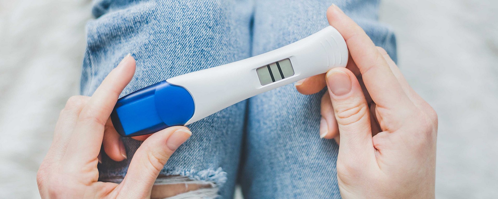 Hände, die einen Schwangerschaftstest mit zwei Strichen – was auf ein positives Ergebnis hindeutet – über den mit Jeans bekleideten Knien halten.