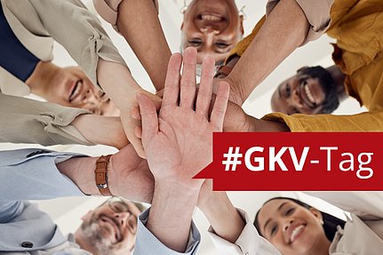 Eine Gruppe von Menschen steht lächelnd im Kreis und hält sich an den Händen; im Bild ist ein rotes Transparent mit der Aufschrift #GKV-Tag zu sehen.