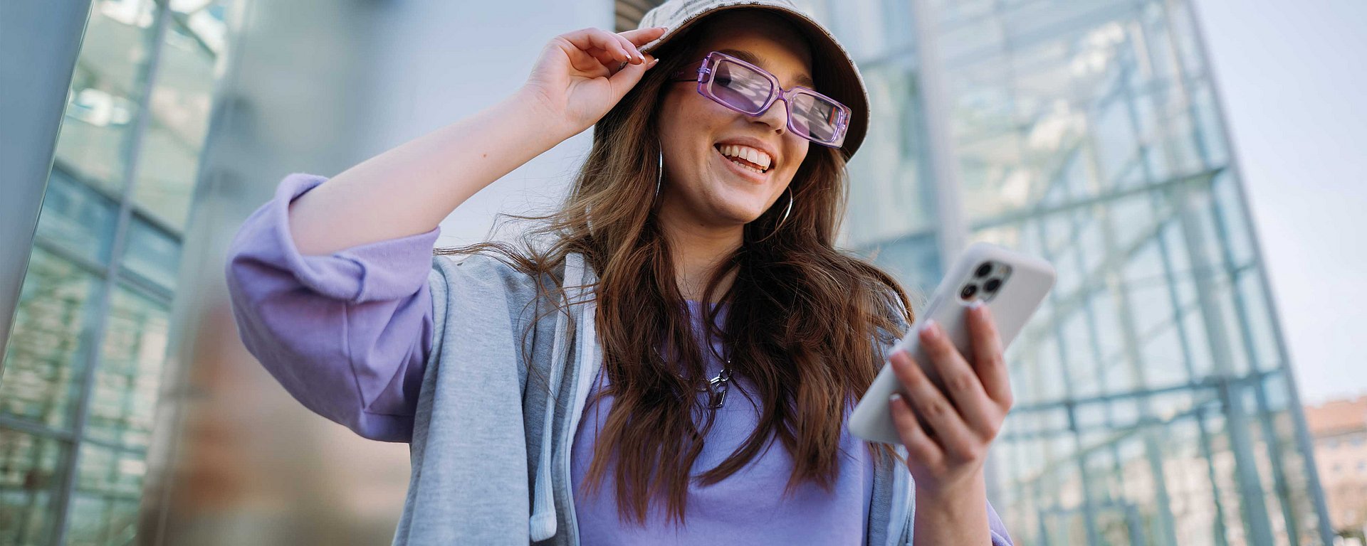 Eine lächelnde Frau mit Hut und Sonnenbrille schaut im Freien in einem modernen, von Glasgebäuden geprägten Bereich auf ihr Handy.