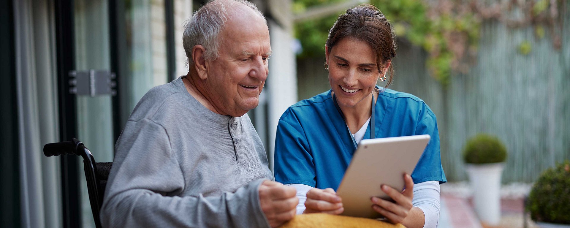 Eine lächelnde Krankenschwester zeigt einem älteren Mann ein Tablet, während sie gemeinsam im Freien sitzen.
