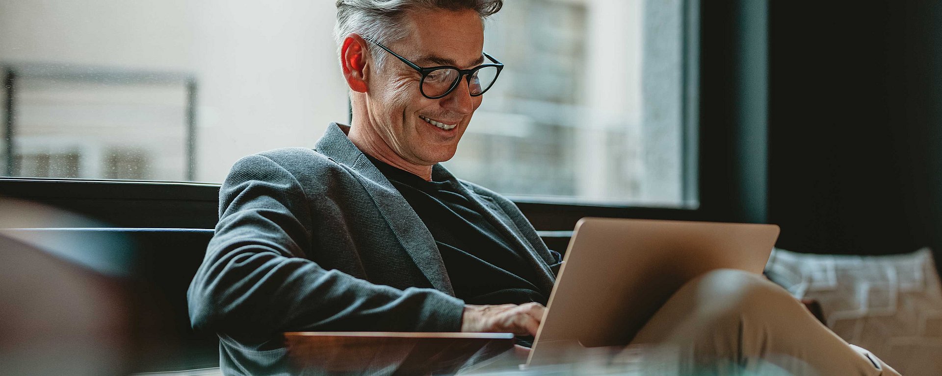 Ein lächelnder Mann mit Brille sitzt drinnen in der Nähe eines großen Fensters und arbeitet an einem Laptop.