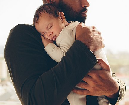 Ein Mann hält tagsüber ein schlafendes Baby sanft in seinen Armen, dicht an seiner Brust.