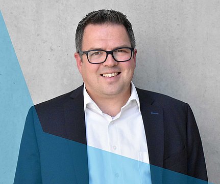 Ein lächelnder Mann mit Brille und Anzugjacke steht vor einer grauen Wand mit einem blauen geometrischen Muster.