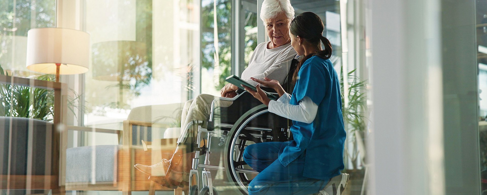 Eine Krankenschwester kniet neben einer älteren Frau im Rollstuhl und bespricht mit ihr etwas auf einem Tablet in einem hellen Raum.