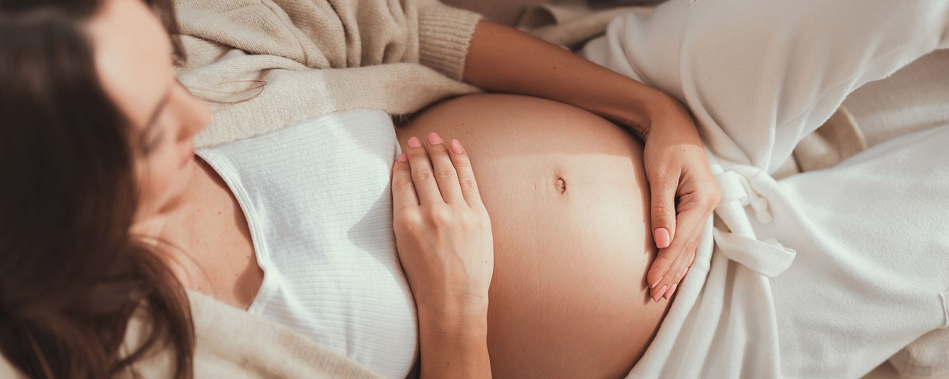 Eine schwangere Frau in weißer Kleidung, die sanft ihren nackten Bauch berührt, während sie sich hinlegt und entspannt.