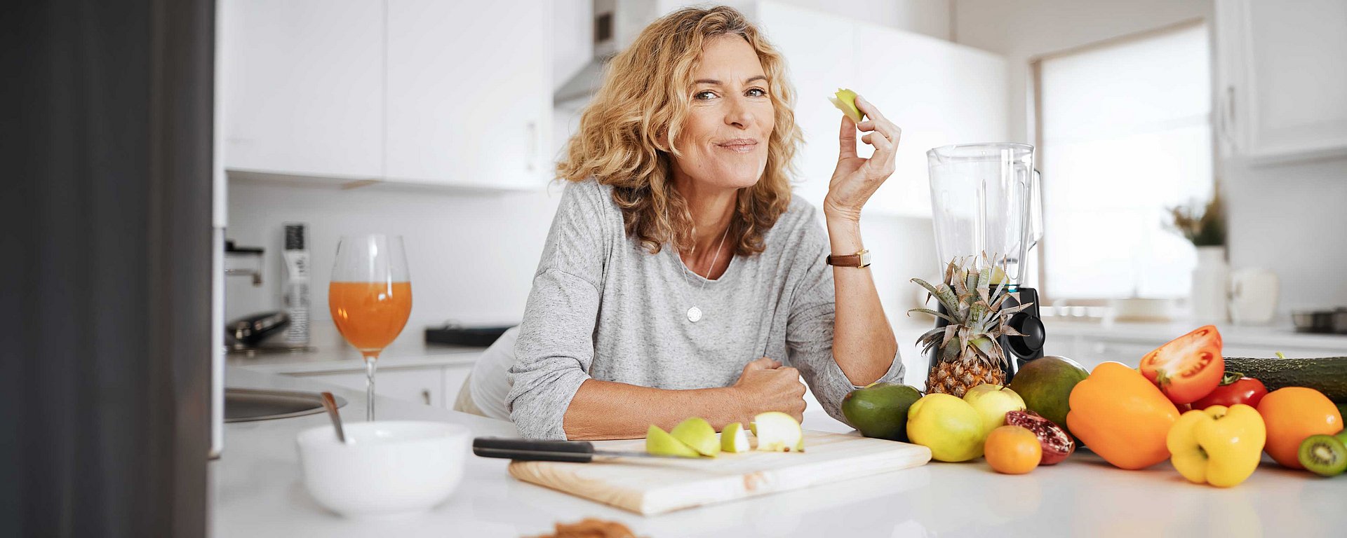 Eine Frau in einer Küche, die Obst isst; vor ihr stehen verschiedene Obstsorten und ein Mixer auf der Arbeitsplatte.
