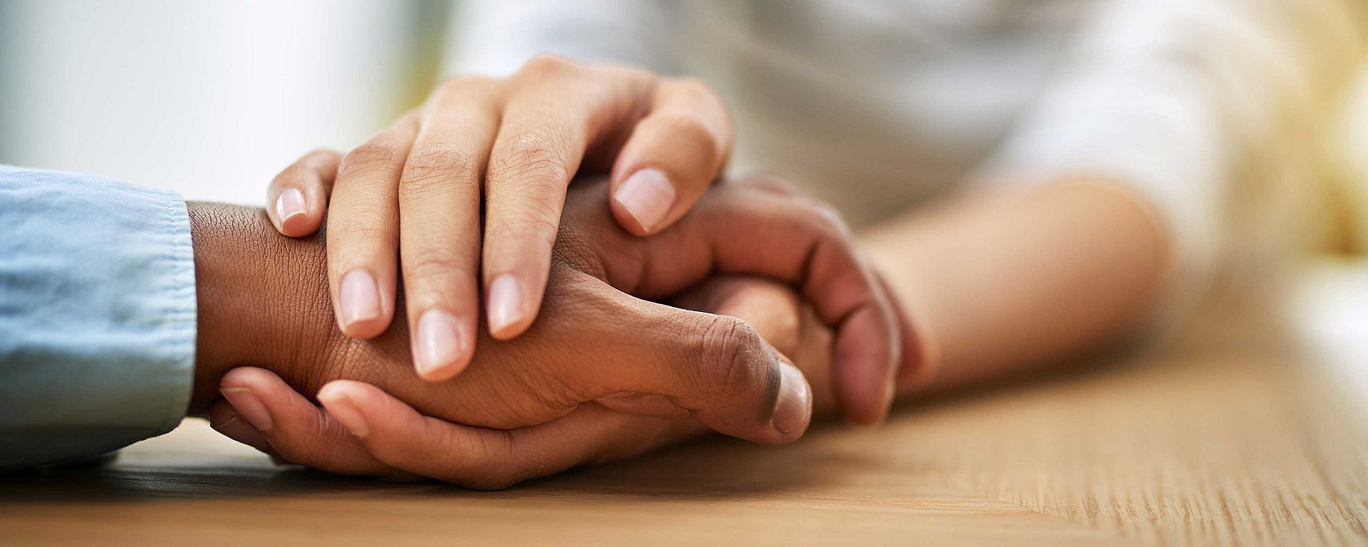Zwei Hände, die in einer tröstenden Geste sanft eine andere Hand auf einer Holzfläche umschließen.