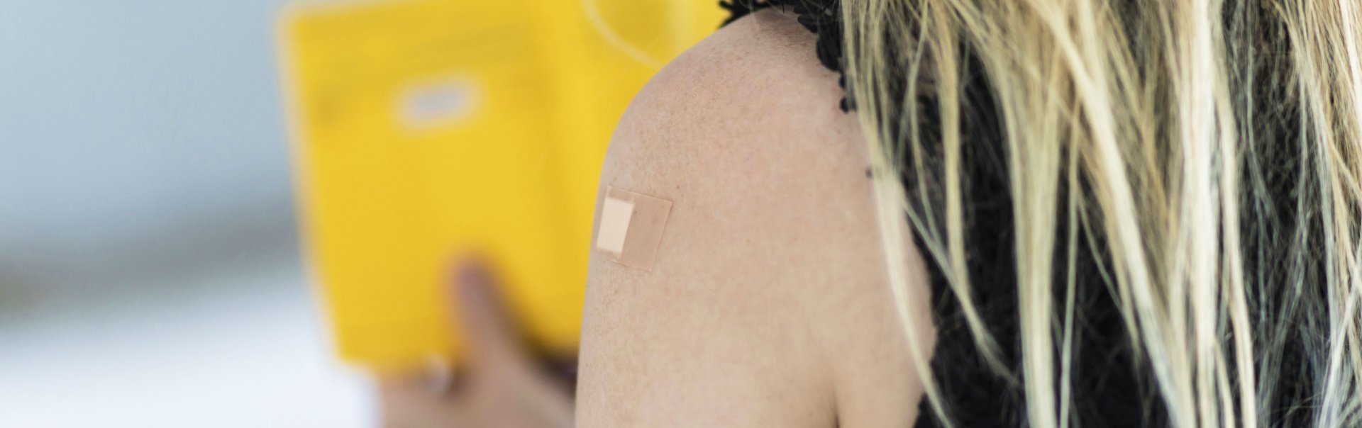 Eine Frau steht mit dem Rücken zur Kamera, hat ein Pflaster von einer Impfung auf dem Arm und hält den Impfpass fest.
