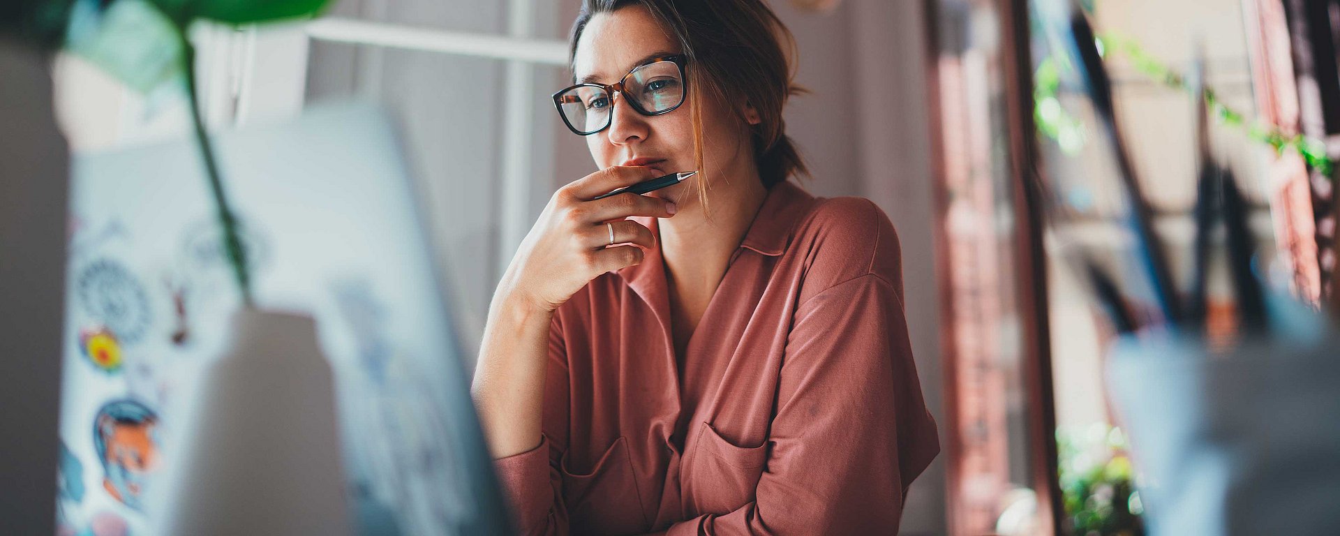 Eine Frau mit Brille und rosa Shirt, die nachdenklich auf den Bildschirm eines Laptops blickt und einen Stift an die Lippen hält.