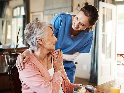 Eine Krankenschwester in blauer Arbeitskleidung lächelt und unterhält sich mit einer älteren Frau, die an einem Tisch sitzt und gerade ein Dessert isst.