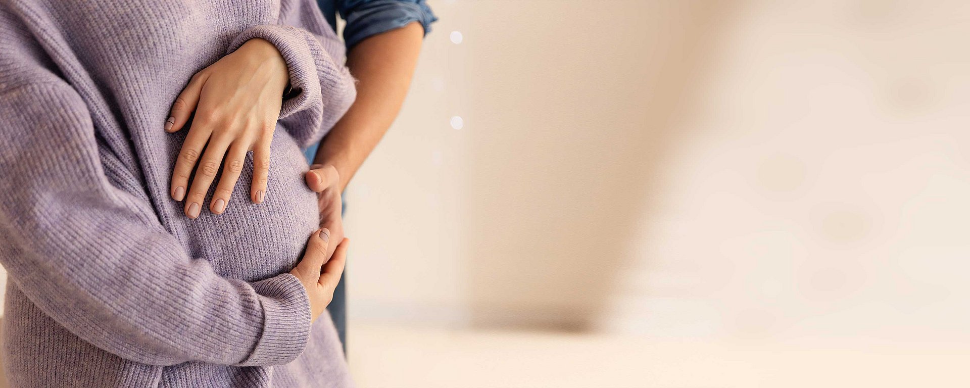 Eine schwangere Person in einem Pullover, deren Hände sanft auf ihrem Bauch ruhen, und eine weitere Person, deren Hand sich dazu gesellt.