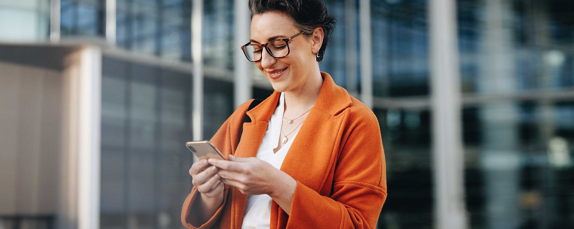 Lächelnde Frau in einem orangefarbenen Mantel, die draußen in einer städtischen Umgebung ihr Smartphone benutzt.