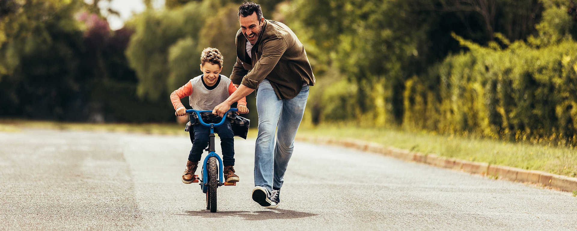 Ein Mann hilft einem kleinen Jungen auf einer ruhigen Straße beim Fahrradfahren; beide lächeln und genießen den Moment.
