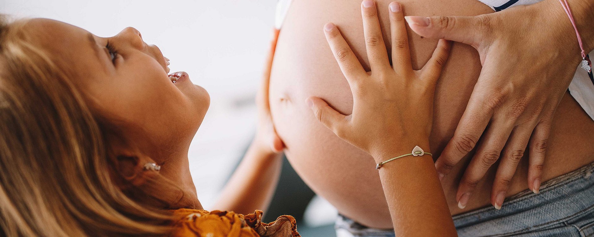 Ein lächelndes Mädchen, das mit beiden Händen den nackten Bauch einer schwangeren Frau berührt.