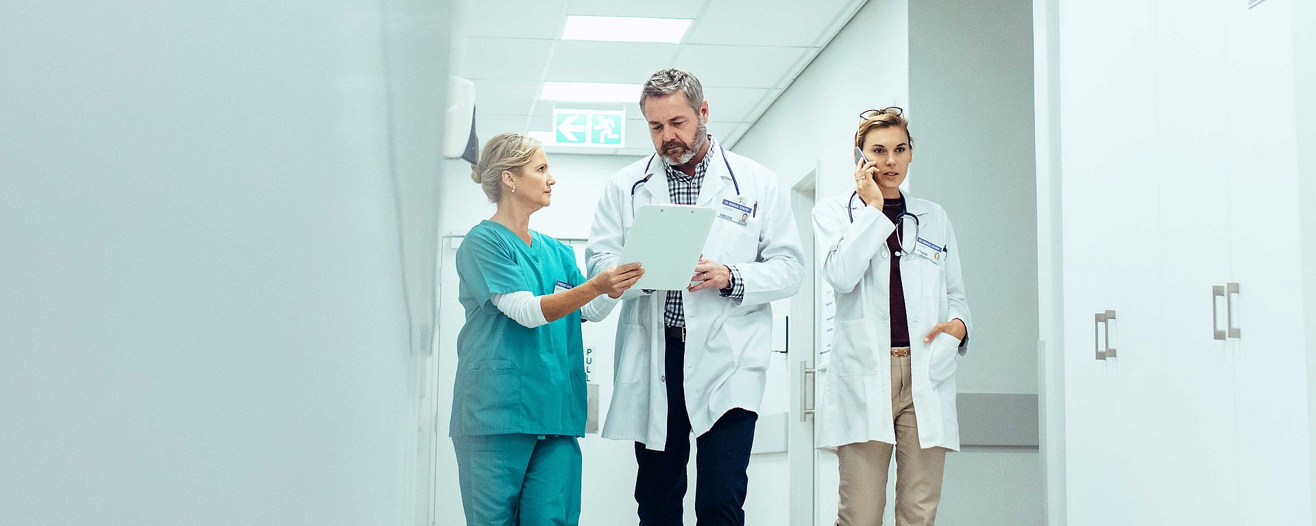 Drei medizinische Fachkräfte gehen durch einen Krankenhausflur, besprechen Unterlagen und telefonieren.