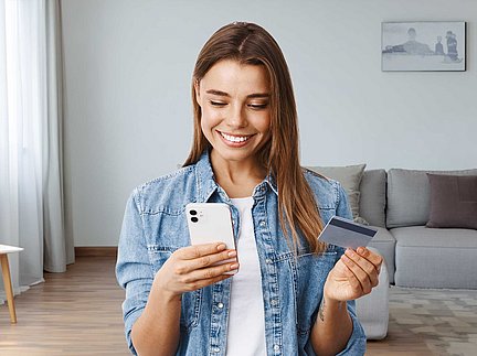 Eine lächelnde Frau sitzt in einem hellen Wohnzimmer und hält ein Smartphone und eine Kreditkarte in der Hand.