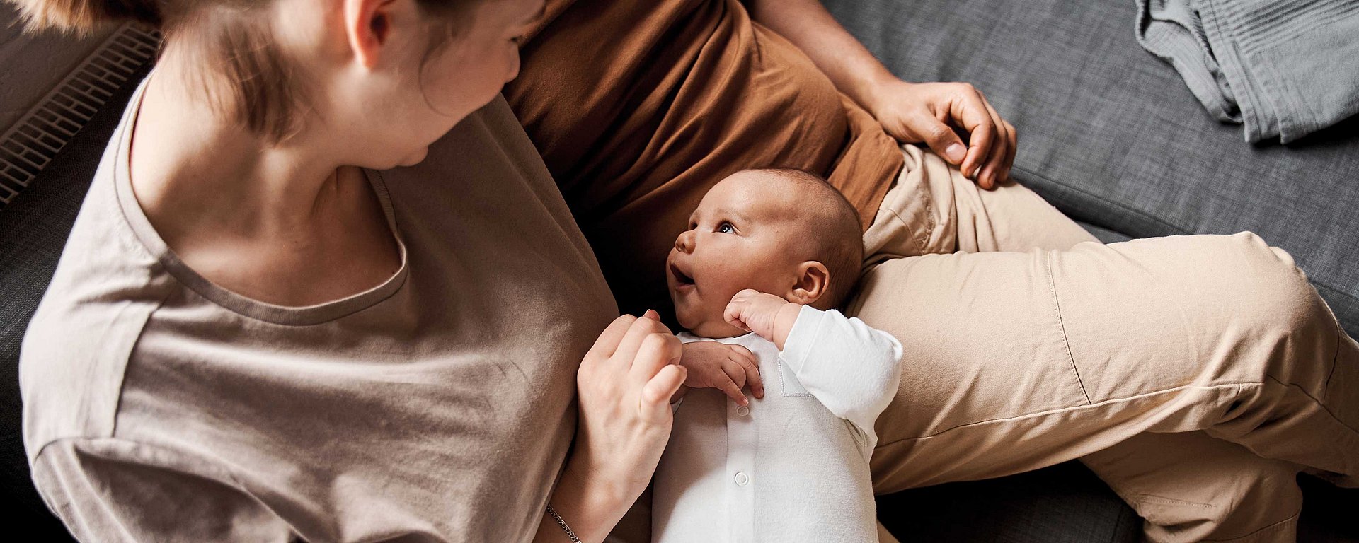 Ein Baby in einem weißen Strampler liegt auf dem Schoß eines Elternteils und blickt zu einem anderen lächelnden Elternteil auf, der neben ihm auf dem Sofa sitzt.