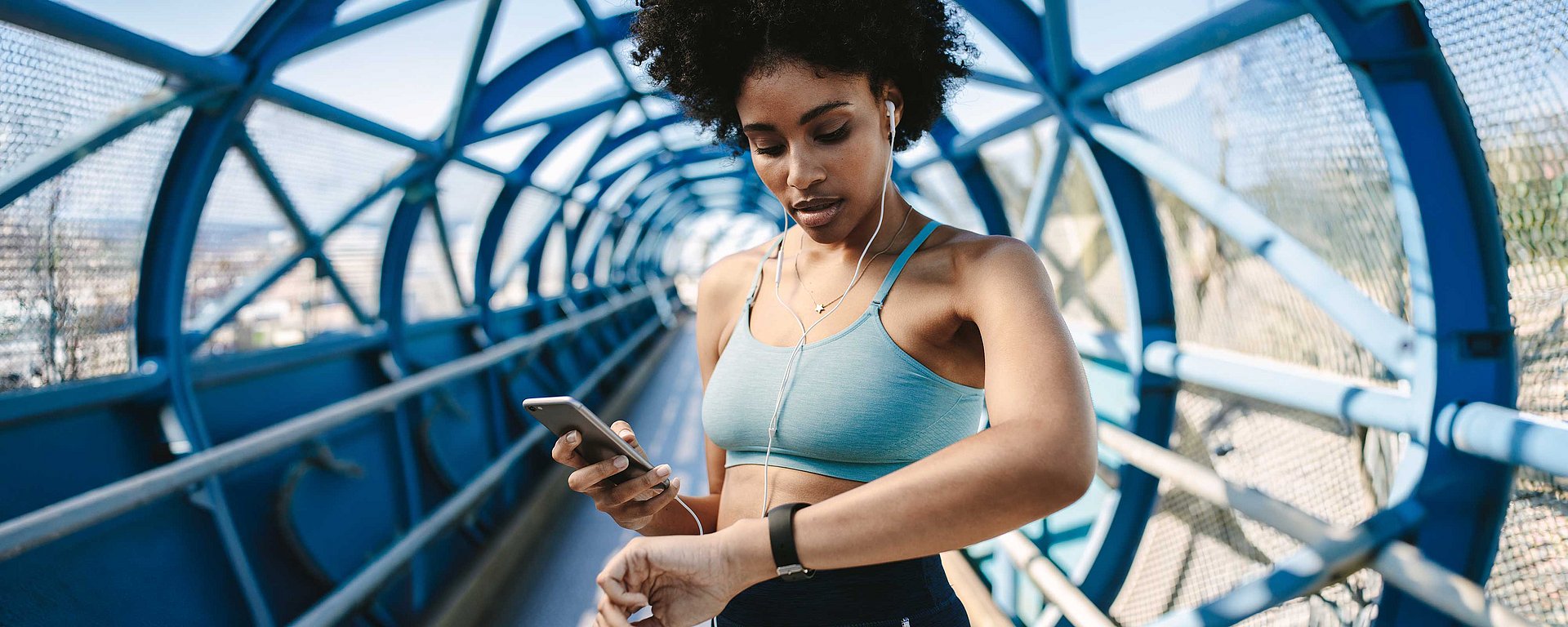 Eine Frau in Sportkleidung steht auf einer blauen Brücke und überprüft ihren Fitness-Tracker, während sie ein Smartphone in der Hand hält.