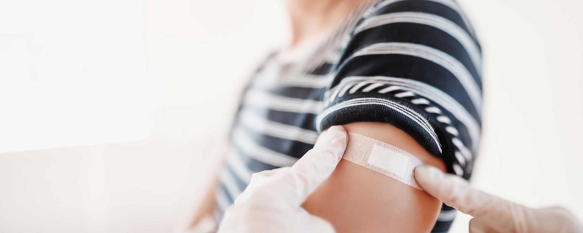 Mit Handschuhen bekleidete Hände legen nach einer Impfung einen Verband am Arm einer Person an.