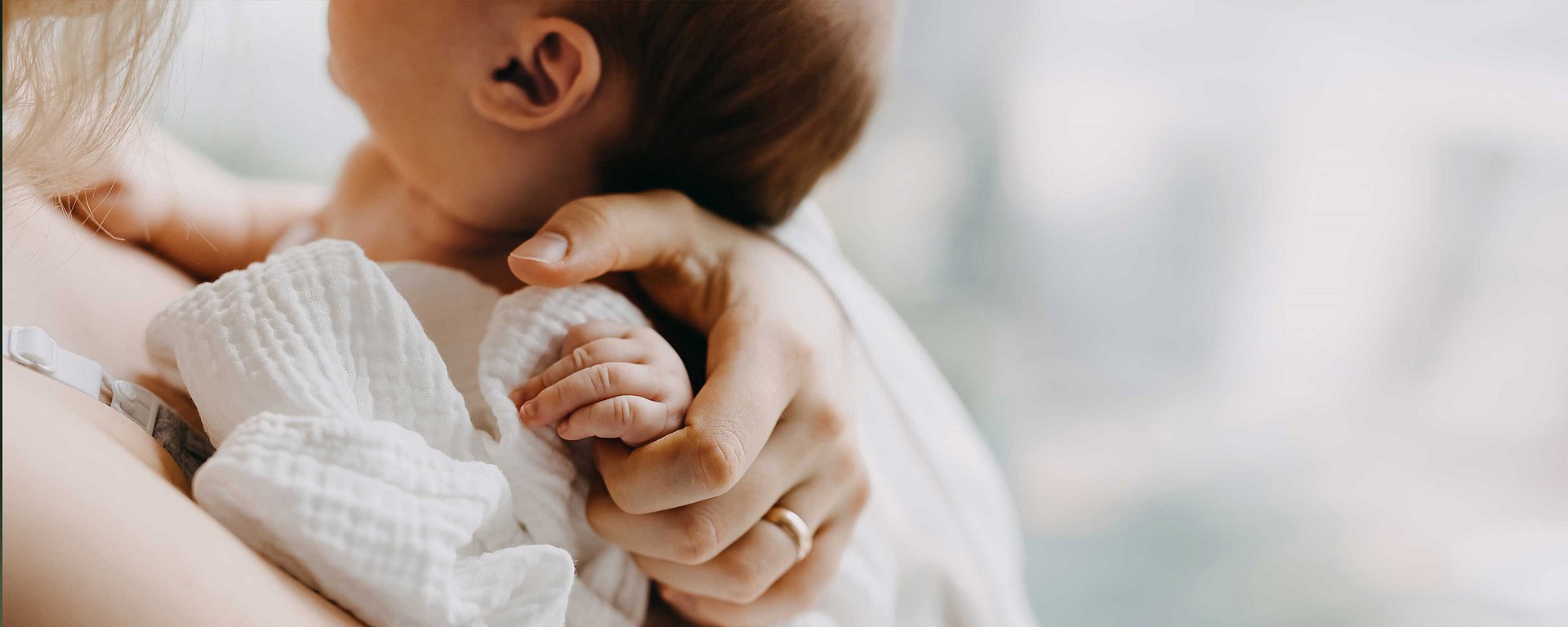 Nahaufnahme einer Frau, die ein in eine weiße Decke gewickeltes Baby im Arm hält, wobei die Hand des Babys auf ihrem Arm ruht.