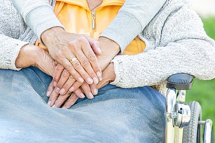 Nahaufnahme der Hände zweier Menschen, die nebeneinander auf dem Schoß ruhen; einer sitzt im Rollstuhl, im Freien.