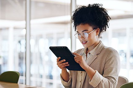 Eine Frau mit Brille sitzt an einem Schreibtisch in einem hellen Büro und nutzt ein Tablet.