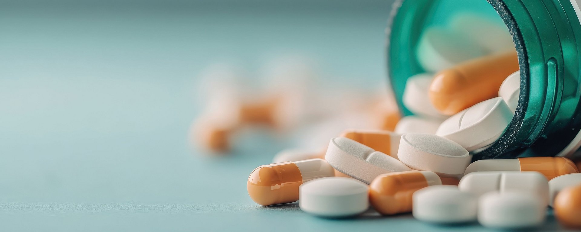 Eine umgekippte grüne Flasche, aus der weiße und orangefarbene Tabletten und Kapseln auf eine blaue Fläche herausfallen.