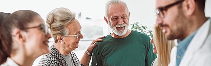 Eine Gruppe lächelnder Erwachsener, darunter Senioren und Ärzte, steht zusammen in einem hellen Raum und unterhält sich.