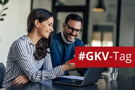 Zwei Menschen, die in die Kamera lächeln, während sie auf einen Laptop blicken; über das Bild verläuft ein rotes Banner mit dem Hashtag #GKV.