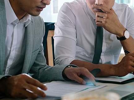 Zwei Männer in Geschäftskleidung, die an einem Schreibtisch Dokumente besprechen; einer hält einen Stift in der Hand, beide sind konzentriert und vertieft in das Gespräch.