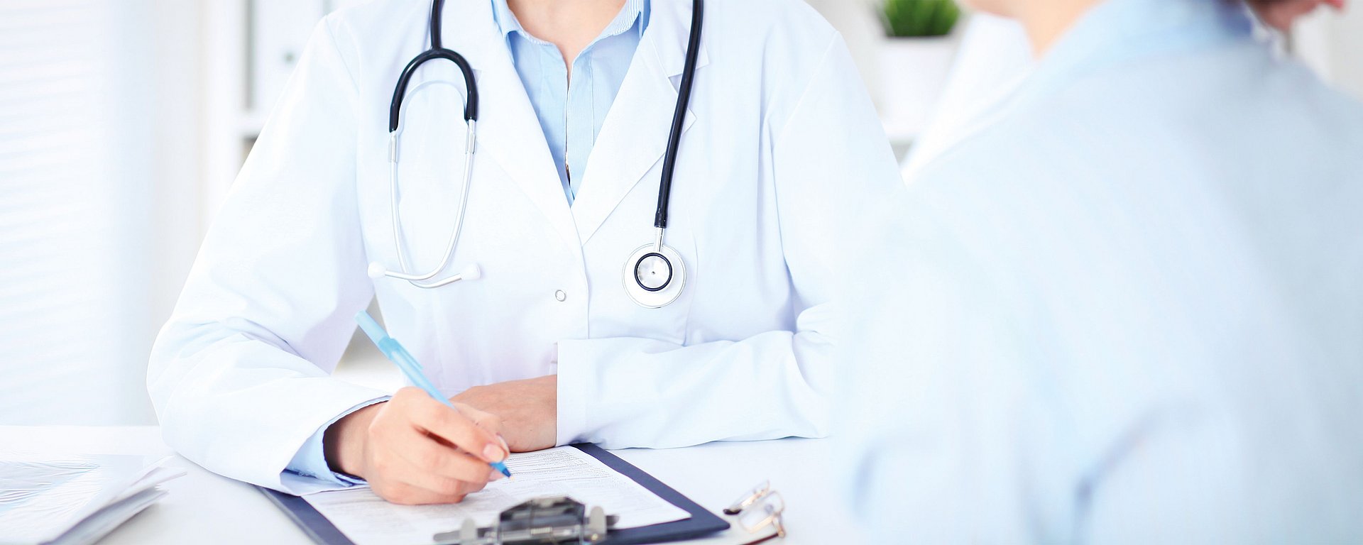 Ein Arzt mit einem Stethoskop, der auf einem Schreibblock notiert, während er mit einem Patienten auf der anderen Seite des Schreibtisches spricht.