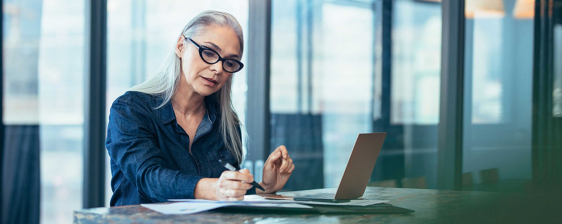 Eine Frau mit grauen Haaren und Brille, die an einem Schreibtisch mit einem Laptop in einem modernen Büro auf ein Blatt Papier schreibt.