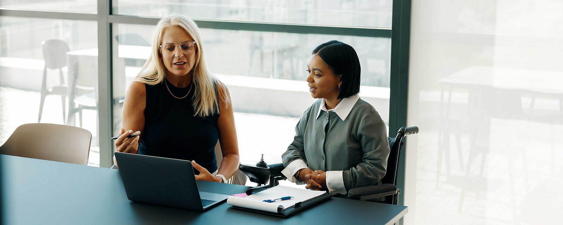 Zwei Frauen unterhalten sich an einem Schreibtisch, auf dem ein Laptop und ein Notizbuch liegen, in einem hellen Büro.