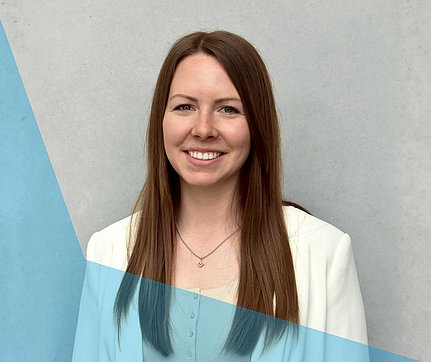 Lächelnde Frau mit langen braunen Haaren in einem weißen Blazer, die vor einem hellgrauen Hintergrund mit blauen Akzenten steht.
