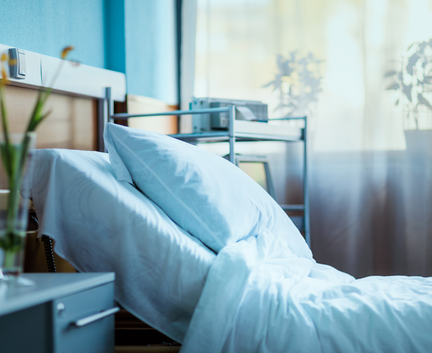 Ein ordentlich gemachtes Krankenhausbett mit weißer Bettwäsche in einem hellen Raum, in den Sonnenlicht fällt und in dem Pflanzen am Fenster stehen.
