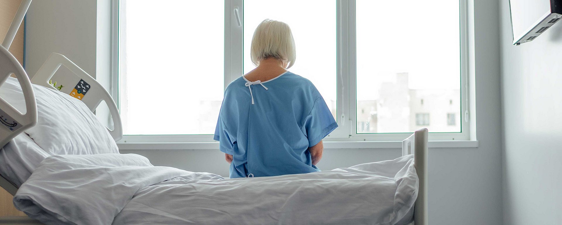 Eine ältere Frau in einem Krankenhauskittel sitzt auf einem Bett und blickt aus einem hellen Fenster in einem Krankenhauszimmer.