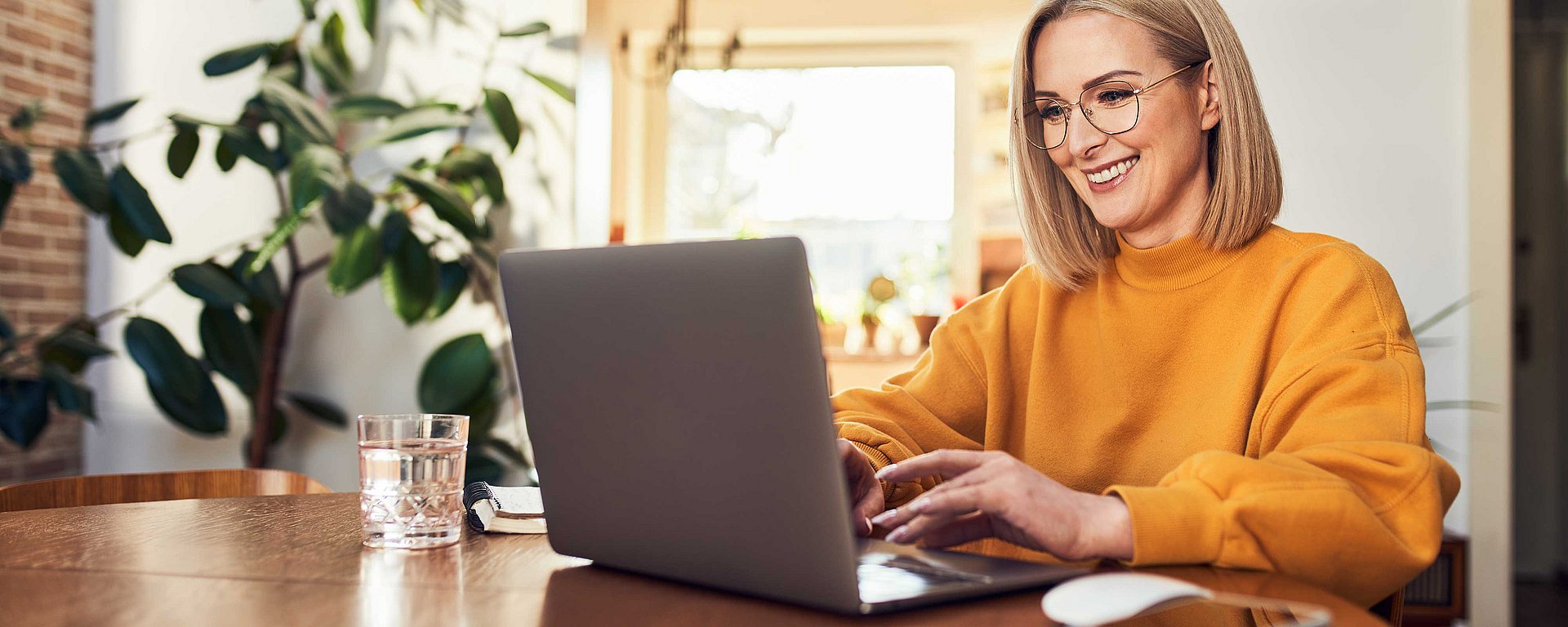 Eine lächelnde Frau mit Brille, die an einem Holztisch in einem hellen Raum an einem Laptop arbeitet, daneben steht ein Glas Wasser.