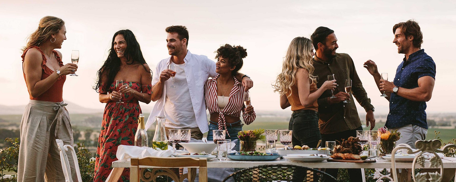 Sieben Menschen stehen im Freien, lächeln und unterhalten sich, während sie ihre Getränke in der Hand halten und um einen festlich gedeckten Tisch herumstehen, auf dem Essen, Gläser und Blumen stehen. Im Hintergrund ist eine malerische Aussicht auf Felder bei Sonnenuntergang zu sehen.
