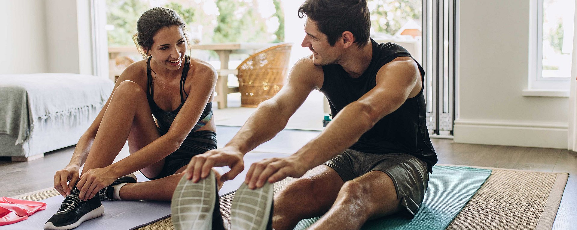 Ein Mann und eine Frau in Sportkleidung, die in einem hellen Raum lächelnd gemeinsam auf Yogamatten Dehnübungen machen.