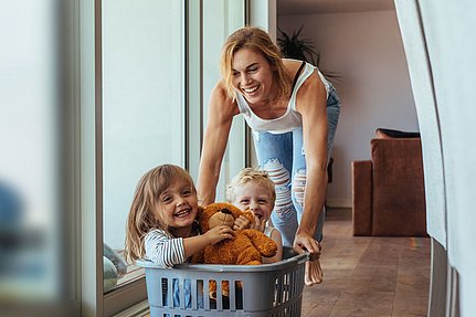 Eine lächelnde Frau schiebt zwei fröhliche Kinder in einem Wäschekorb einen Flur entlang.