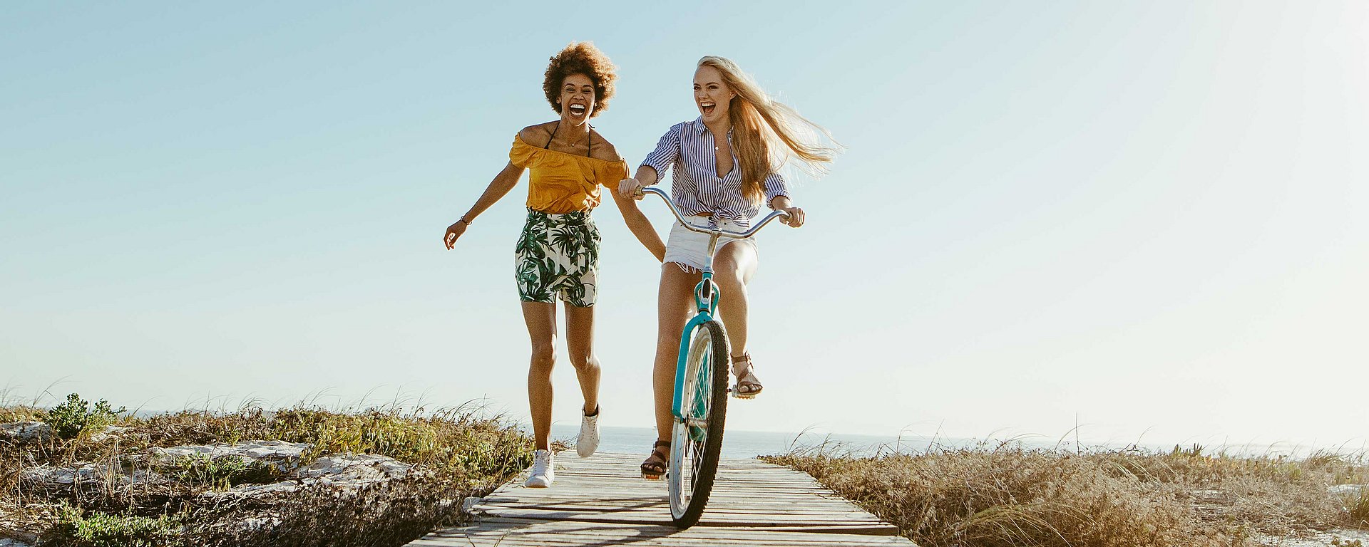 Zwei Frauen genießen einen sonnigen Tag: Die eine fährt Fahrrad, die andere läuft neben ihr auf einer Promenade am Meer entlang.